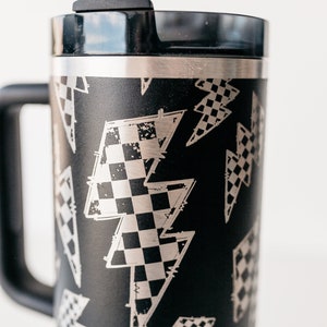 Op de afbeelding: Zwarte reismok met een handvat en een deksel, met een patroon van zilveren bliksemschichten met een dambordontwerp. Inclusief een zilveren rietje. De mok is ontworpen voor warme of koude dranken.