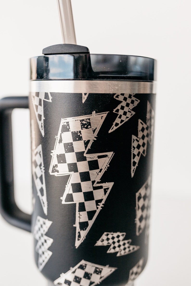 Op de afbeelding: Zwarte reismok met een handvat en een deksel, met een patroon van zilveren bliksemschichten met een dambordontwerp. Inclusief een zilveren rietje. De mok is ontworpen voor warme of koude dranken.