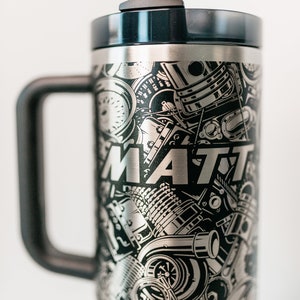May include: Stainless steel tumbler with a black handle and lid. The tumbler is covered in a black and silver engraved design of engine parts and the name "MATT". Includes a clear straw.