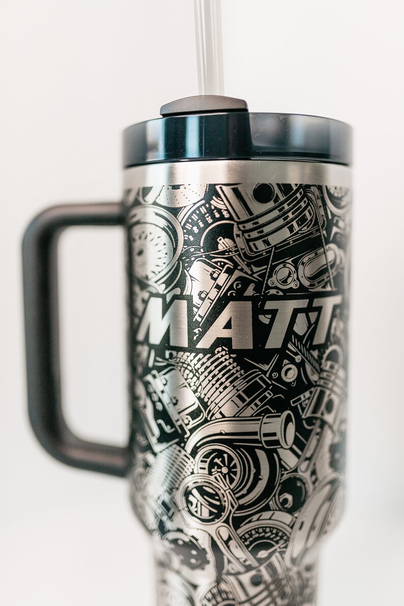 May include: Stainless steel tumbler with a black handle and lid. The tumbler is covered in a black and silver engraved design of engine parts and the name "MATT". Includes a clear straw.