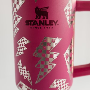 Op de afbeelding: Een magenta Stanley-beker met een zilveren bliksem- en geblokt vlagpatroon. De beker heeft een handvat en een zilveren rand. Het Stanley-logo en "SINCE 1913" staan op de voorkant.