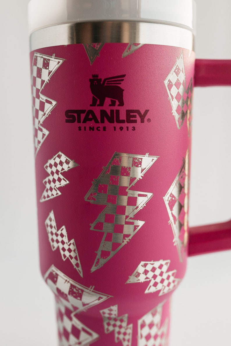 Op de afbeelding: Een magenta Stanley-beker met een zilveren bliksem- en geblokt vlagpatroon. De beker heeft een handvat en een zilveren rand. Het Stanley-logo en "SINCE 1913" staan op de voorkant.