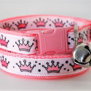 May include: Pink cat collar with a white background and pink crowns. The collar has a pink buckle and a silver bell.