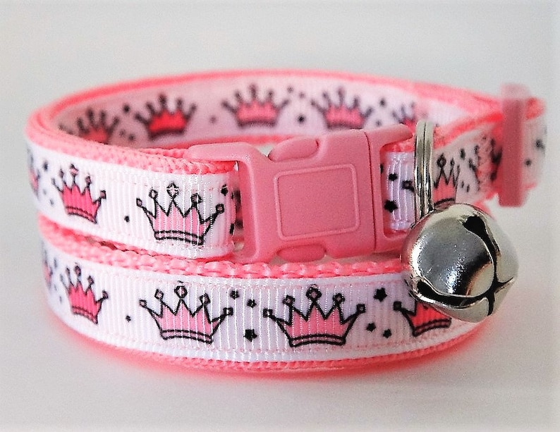 May include: Pink cat collar with a white background and pink crowns. The collar has a pink buckle and a silver bell.