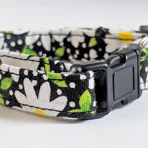 May include: A black and white cat collar with a daisy print and green leaves. The collar has a black plastic buckle and a silver bell.