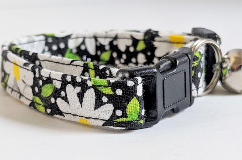 May include: A black and white cat collar with a daisy print and green leaves. The collar has a black plastic buckle and a silver bell.