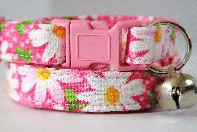 May include: Pink cat collar with a white daisy print and a silver bell. The collar has a pink plastic buckle.