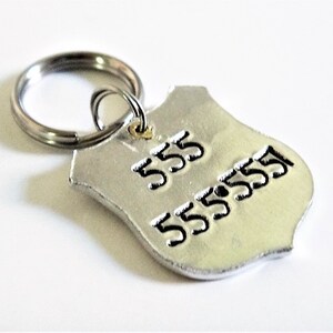 May include: A silver-colored, shield-shaped pet tag with the numbers "555" and "5555551" stamped on it. The tag is attached to a silver split ring. The tag is likely for a dog or cat.
