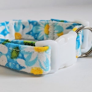 May include: A white cat collar with a blue and yellow floral pattern, a white plastic buckle, and a silver bell.