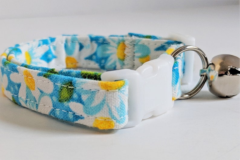 May include: A white cat collar with a blue and yellow floral pattern, a white plastic buckle, and a silver bell.