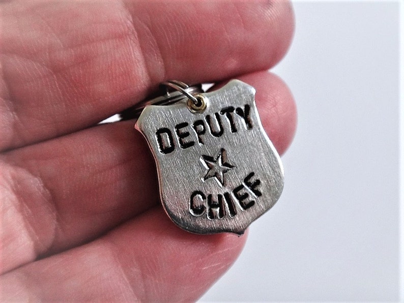 May include: A silver-colored Deputy Chief badge keychain. The badge is shield-shaped with the words "DEPUTY CHIEF" and a star emblem. The keychain is held by a person.