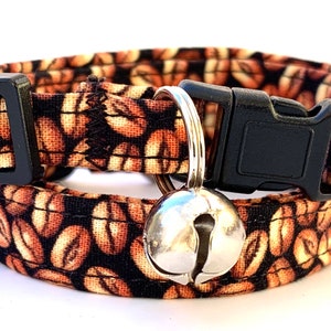 May include: A brown and black cat collar with a coffee bean pattern and a silver bell. The collar has a black plastic buckle.