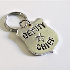May include: A silver-colored keychain featuring a shield-shaped tag. The tag has the words "DEPUTY CHIEF" and a star emblem. The keychain ring is silver and attached to the tag with a small gold-colored ring.