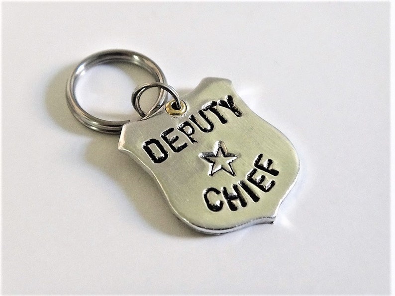 May include: A silver-colored keychain featuring a shield-shaped tag. The tag has the words "DEPUTY CHIEF" and a star emblem. The keychain ring is silver and attached to the tag with a small gold-colored ring.