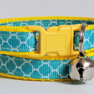 May include: A turquoise and yellow cat collar with a geometric pattern and a silver bell. The collar has a yellow plastic buckle.