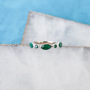 May include: A silver ring with five emerald green gemstones set in a row. The ring is on a white marble surface.