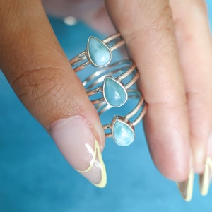 May include: A set of four silver rings with teardrop-shaped blue gemstone accents. The rings are stacked on each other and are being held by a hand.