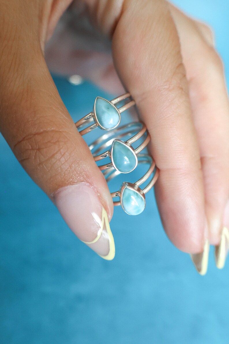 May include: A set of four silver rings with teardrop-shaped blue gemstone accents. The rings are stacked on each other and are being held by a hand.