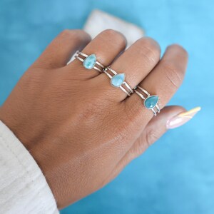 May include: Three silver rings with teardrop-shaped blue gemstone accents. The rings are stacked on a person's finger.