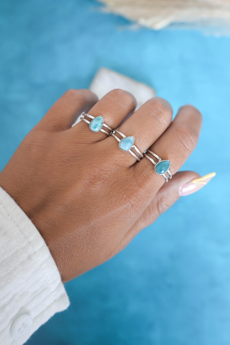 May include: Three silver rings with teardrop-shaped blue gemstone accents. The rings are stacked on a person's finger.