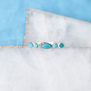 May include: A silver ring with five turquoise stones. The ring has an oval central stone and four smaller, round stones. The ring is displayed on a white and blue surface.