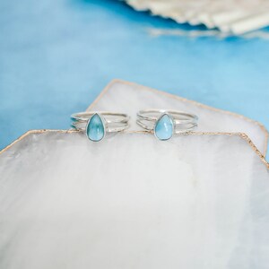 May include: Two silver rings with teardrop-shaped blue gemstones on a white background.