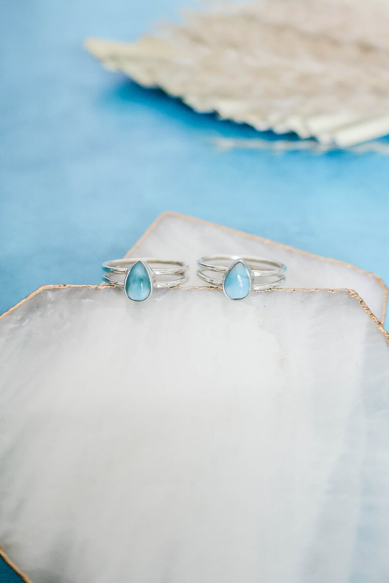 May include: Two silver rings with teardrop-shaped blue gemstones on a white background.