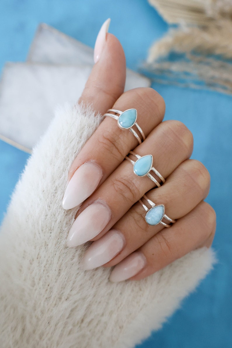 May include: Three silver rings with teardrop-shaped turquoise gemstones. The rings are stacked on a finger with light pink nail polish.