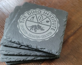 Puede incluir: Conjunto de cuatro posavasos de pizarra negra con un logotipo grabado en gris que dice "The Dino Institute" con un dinosaurio y otros símbolos.