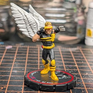 Puede incluir: Figura en miniatura de superhéroe con alas blancas, visera amarilla y botas amarillas. La figura lleva un traje negro y amarillo y está sobre una base circular negra con detalles rojos y blancos. Un artículo de colección.