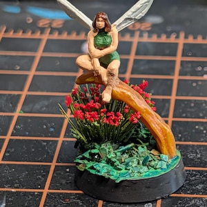 May include: A miniature figurine of a winged figure with brown hair wearing a green outfit, sitting on a curved brown branch. The base features a cluster of small red flowers and green foliage, all mounted on a circular black pedestal.