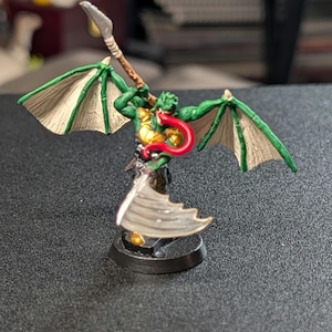 May include: A painted miniature figurine of a winged creature with green skin, a long red tongue, and gold armor. The figure holds a spear in one hand and features large, textured, off-white wings with green edges, standing on a circular black base.