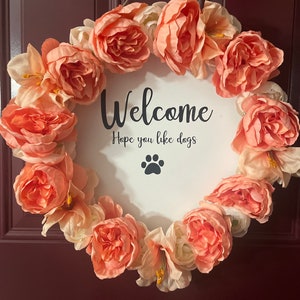Pode incluir: Uma coroa de flores cor de pêssego com uma placa branca que diz "Welcome Hope you like dogs" com uma pegada de pata preta.