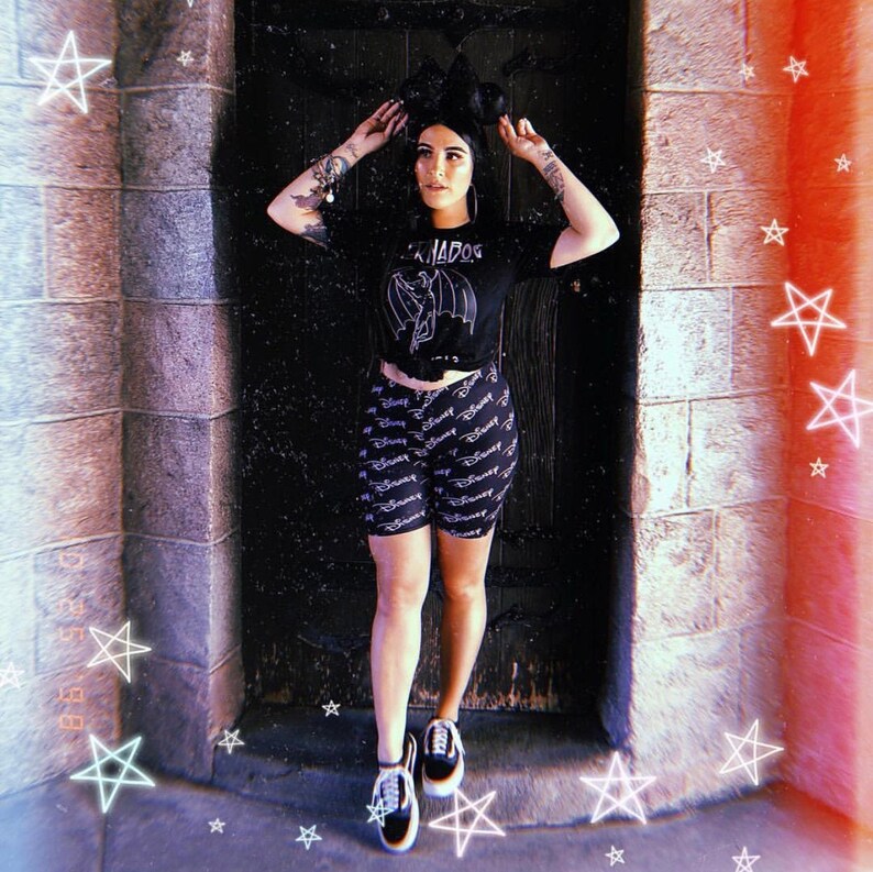 Puede incluir: Una mujer con una camiseta negra con un gr&aacute;fico de un murci&eacute;lago y el texto "Bad Dog" en ella. Tambi&eacute;n lleva pantalones cortos negros y blancos con la palabra "Disney" repetida por todas partes. Est&aacute; de pie frente a una puerta de madera oscura con una pared de piedra detr&aacute;s de ella.