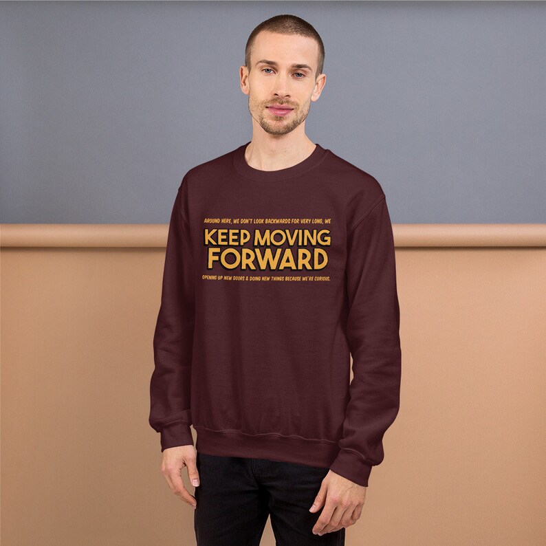 Puede incluir: Una sudadera granate con el texto "KEEP MOVING FORWARD" en letras doradas. La sudadera tambi&eacute;n tiene texto adicional encima y debajo de la frase principal. La persona de la imagen lleva pantalones negros.