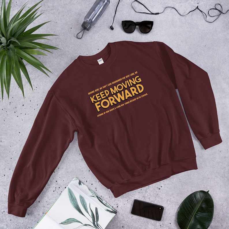 Puede incluir: Una sudadera granate con el texto "KEEP MOVING FORWARD" en amarillo y blanco. La sudadera tiene texto adicional encima y debajo de la frase principal. La prenda se muestra sobre una superficie gris claro.