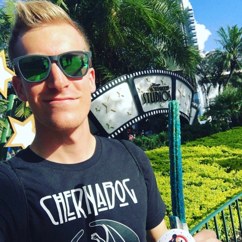 Puede incluir: Un hombre con gafas de sol y una camiseta negra con el texto "Chernabog" impreso en ella. Est&aacute; sosteniendo un churro azul y verde en su mano derecha. El fondo muestra un letrero de Hollywood Studios con un dise&ntilde;o de rollo de pel&iacute;cula.