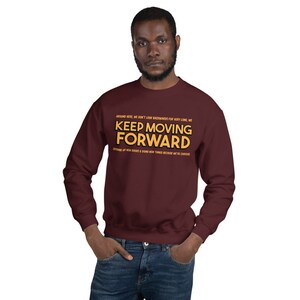 Puede incluir: Una sudadera burdeos con la frase "KEEP MOVING FORWARD" en letras amarillas. La sudadera tambi&eacute;n tiene texto adicional encima y debajo de la frase principal. La persona lleva la sudadera.