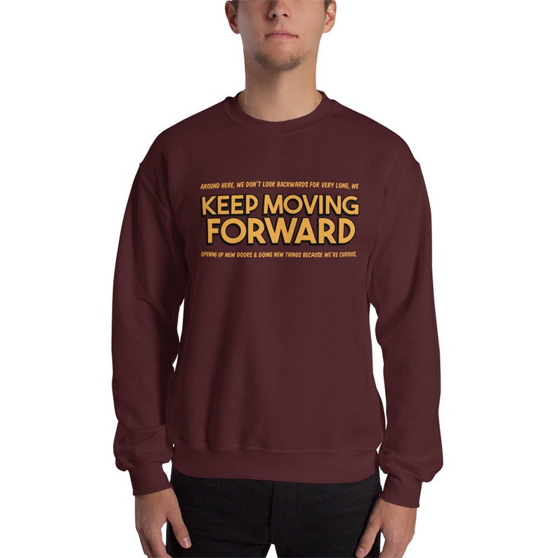Puede incluir: Una sudadera granate con la frase "KEEP MOVING FORWARD" en letras doradas. La sudadera tambi&eacute;n tiene texto adicional encima y debajo de la frase principal. El tema general es motivacional e inspirador.