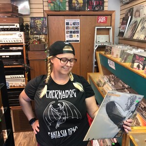 Puede incluir: Una mujer con una camiseta negra con un gr&aacute;fico blanco de una criatura con alas y el texto "Chernados Fantasia 1940" est&aacute; navegando por una tienda de discos. Ella sostiene un disco de vinilo con la portada del &aacute;lbum "Led Zeppelin" en su mano derecha.