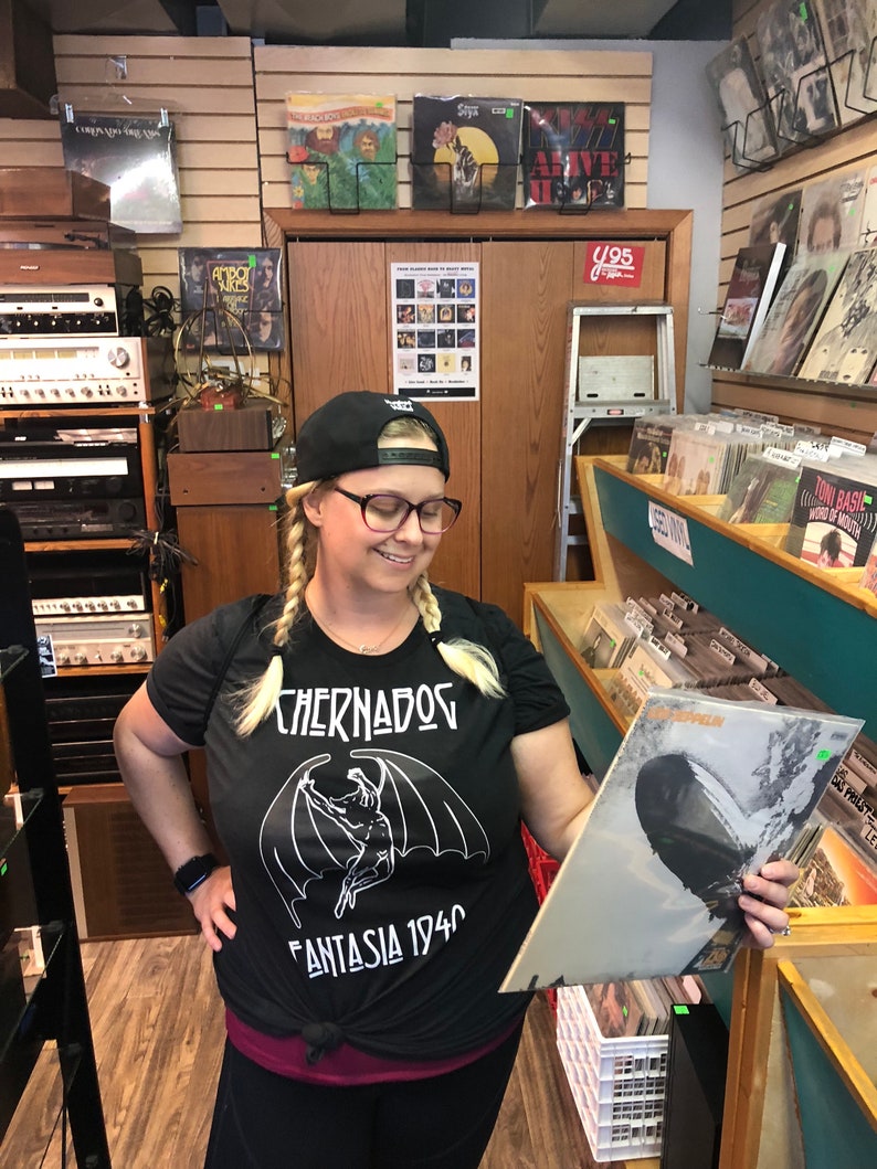 Puede incluir: Una mujer con una camiseta negra con un gr&aacute;fico blanco de una criatura con alas y el texto "Chernados Fantasia 1940" est&aacute; navegando por una tienda de discos. Ella sostiene un disco de vinilo con la portada del &aacute;lbum "Led Zeppelin" en su mano derecha.