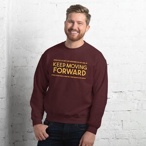 Puede incluir: Una sudadera burdeos con la frase "KEEP MOVING FORWARD" en letras doradas. La sudadera tambi&eacute;n tiene texto adicional encima y debajo de la frase principal. La persona de la imagen est&aacute; sonriendo.