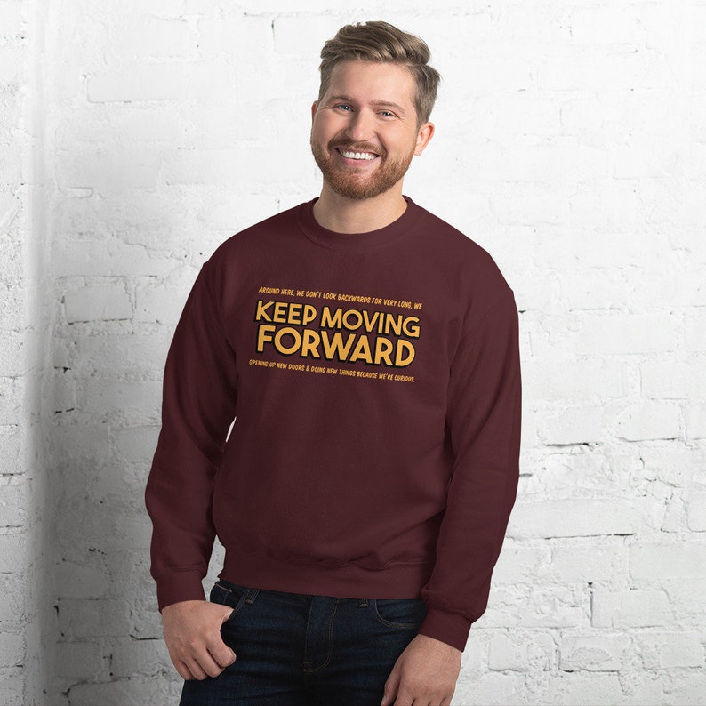 Puede incluir: Una sudadera burdeos con la frase "KEEP MOVING FORWARD" en letras doradas. La sudadera tambi&eacute;n tiene texto adicional encima y debajo de la frase principal. La persona de la imagen est&aacute; sonriendo.