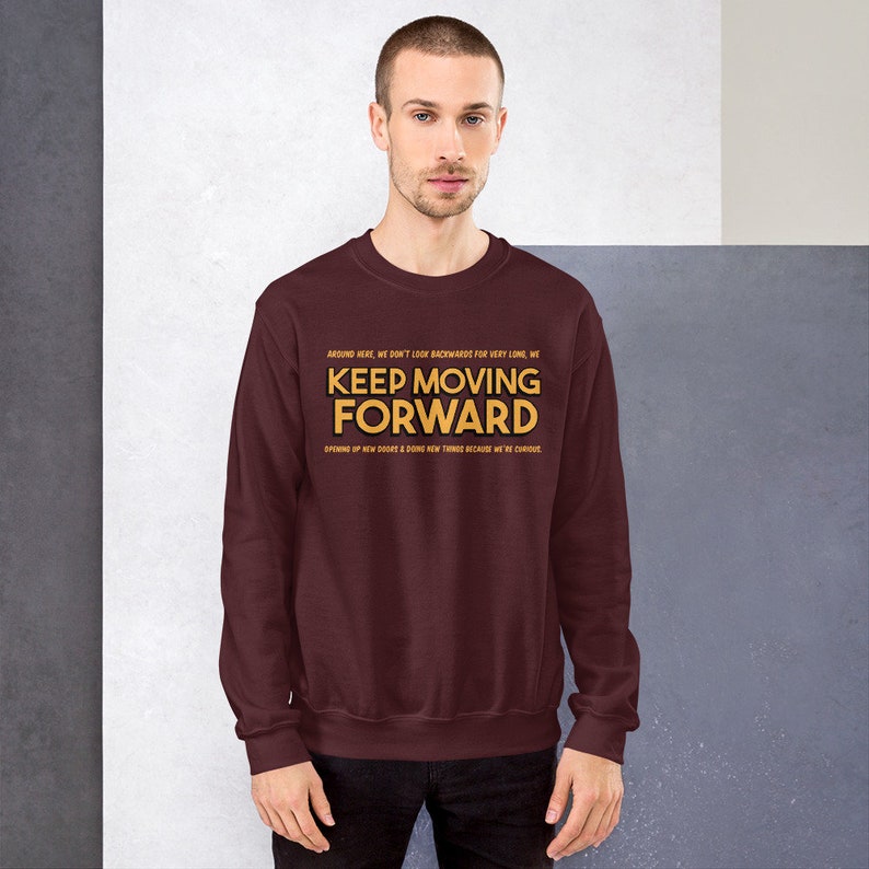 Puede incluir: Una sudadera granate con la frase "KEEP MOVING FORWARD" en letras doradas. La sudadera tambi&eacute;n tiene texto adicional encima y debajo de la frase principal. La persona de la imagen lleva una camisa de color oscuro.