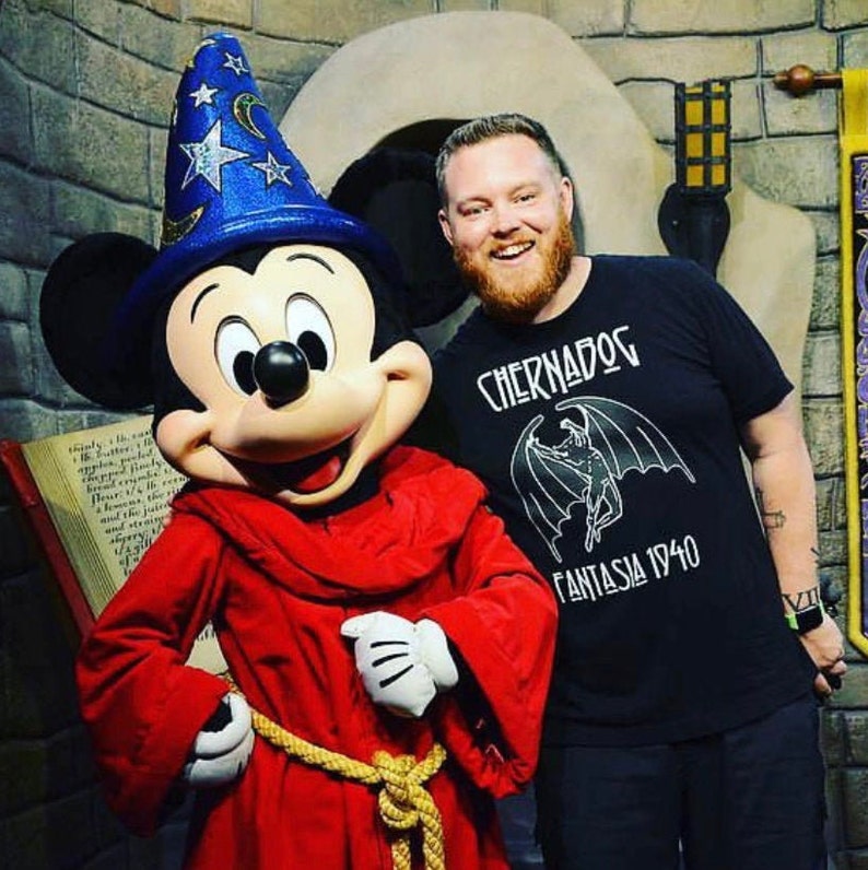 Puede incluir: Mickey Mouse vestido de mago con una t&uacute;nica roja y un sombrero azul con estrellas. Est&aacute; de pie junto a un hombre que lleva una camiseta negra con el texto "Chernabog Fantasia 1940".