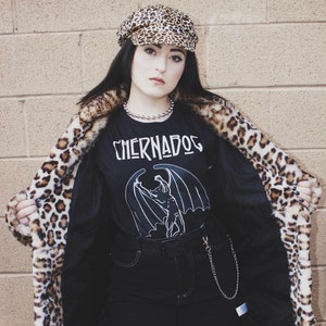 Puede incluir: Una persona lleva una camiseta negra de manga larga con un gr&aacute;fico blanco de una criatura alada y el texto "Chernados". La camiseta se lleva debajo de una chaqueta de piel sint&eacute;tica de leopardo. La persona lleva una gorra de newsboy de leopardo.