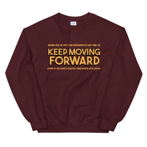 Puede incluir: Una sudadera burdeos con la frase "KEEP MOVING FORWARD" en letras doradas. La sudadera tambi&eacute;n tiene las frases "AROUND HERE, WE DON'T LOOK BACKWARDS FOR VERY LONG, WE" y "OPENING UP NEW DOORS & DOING NEW THINGS BECAUSE WE'RE CURIOUS."