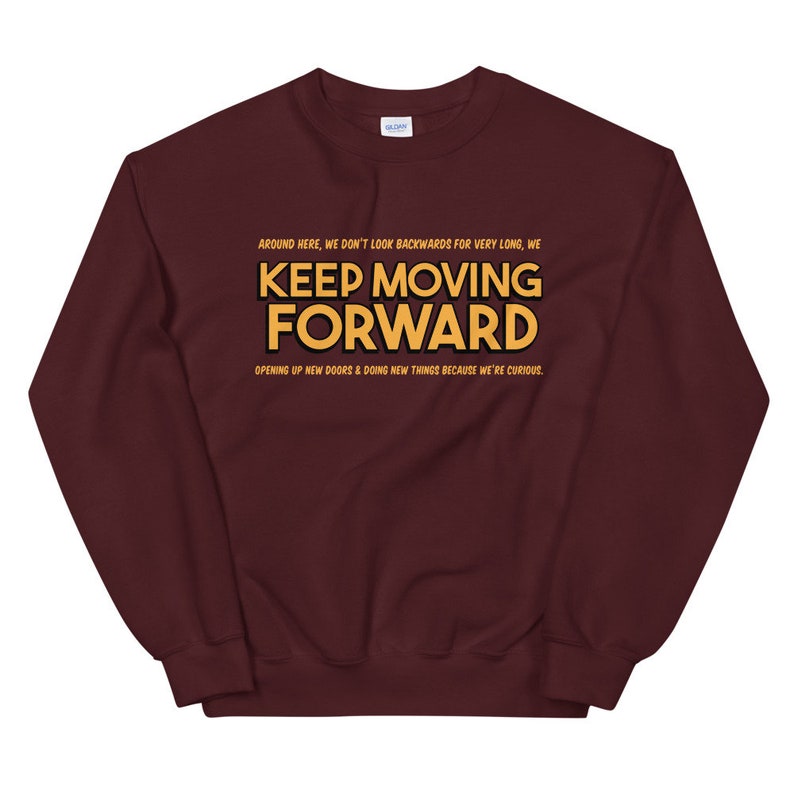 Puede incluir: Una sudadera burdeos con la frase "KEEP MOVING FORWARD" en letras doradas. La sudadera tambi&eacute;n tiene las frases "AROUND HERE, WE DON'T LOOK BACKWARDS FOR VERY LONG, WE" y "OPENING UP NEW DOORS & DOING NEW THINGS BECAUSE WE'RE CURIOUS."