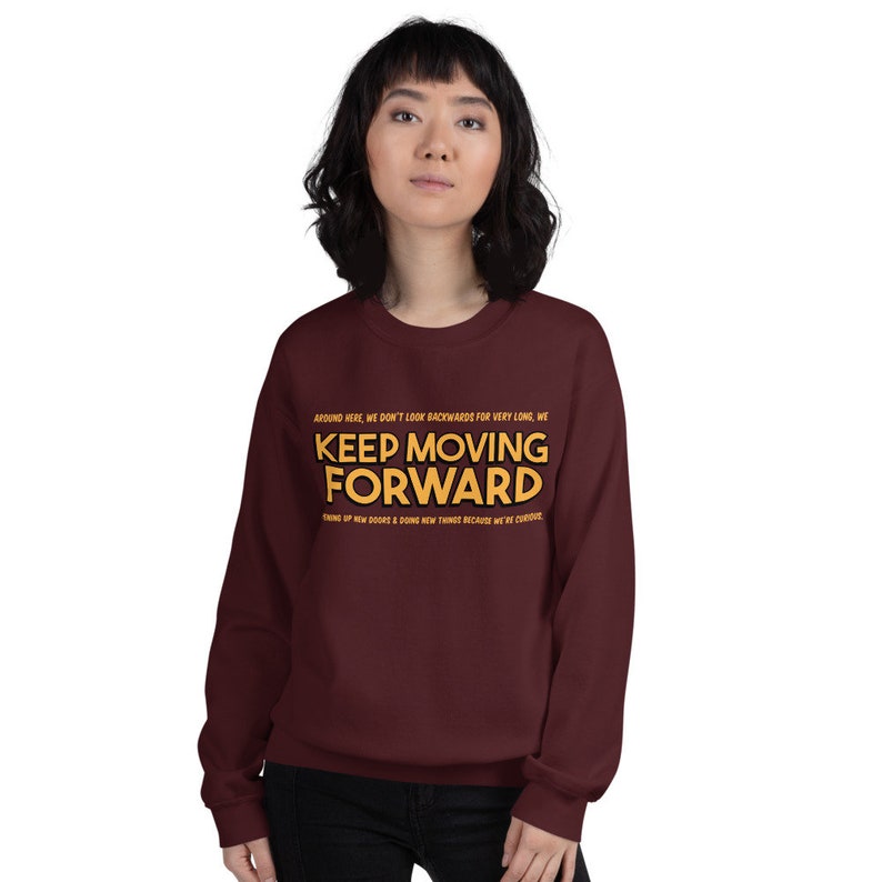 Puede incluir: Una sudadera burdeos con la frase "KEEP MOVING FORWARD" en letras doradas. El texto va acompa&ntilde;ado de texto m&aacute;s peque&ntilde;o arriba y abajo. La sudadera es de cuello redondo y la lleva una persona.