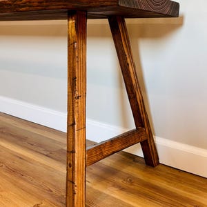 May include: A rustic wooden bench with a dark stained top and light brown legs. The bench has a natural wood grain and a simple, sturdy design. A glass vase with greenery sits on top.